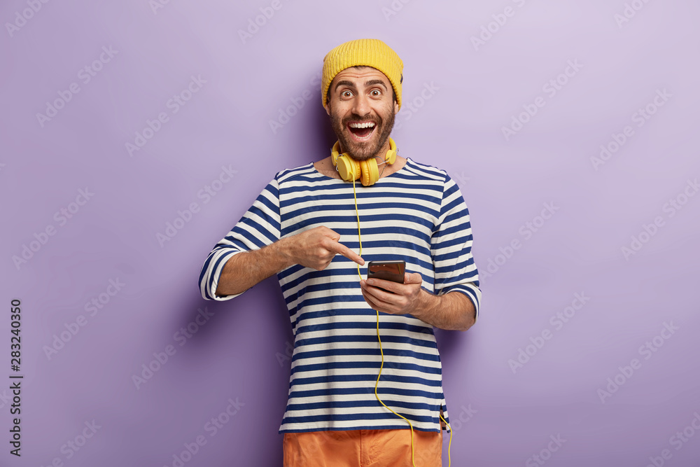 Positive male points into smart phone, glad to get good message, uses cellular and headphones for listening radio online, has bristle, wears yellow hat and striped loose jumper. People and technology