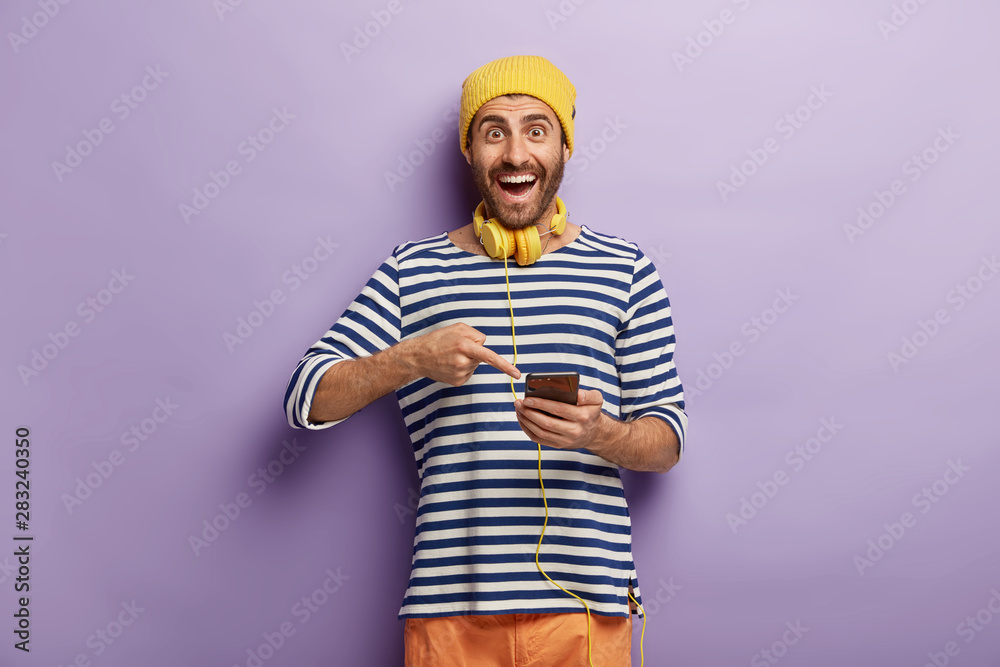 Positive male points into smart phone, glad to get good message, uses cellular and headphones for listening radio online, has bristle, wears yellow hat and striped loose jumper. People and technology Positive male points into smart phone, glad to get good message, uses cellular and headphones for listening radio online, has bristle, wears yellow hat and striped loose jumper. People and technology