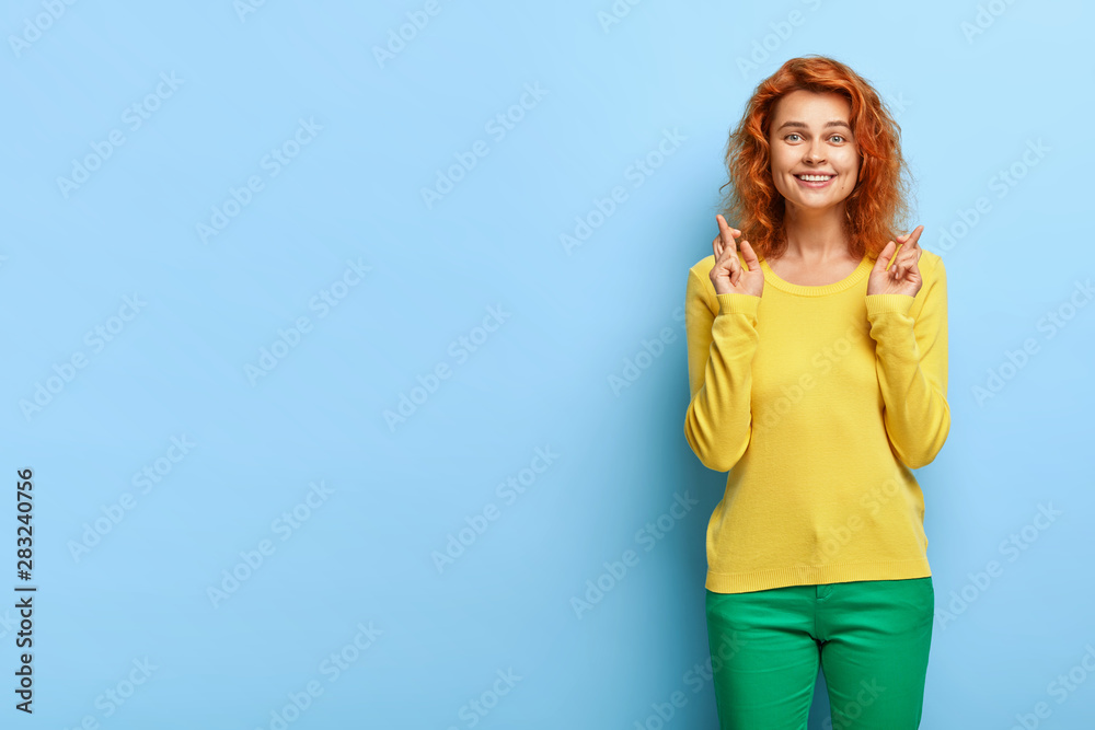 Fototapeta premium Half length shot of glad hopeful ginger female makes wish and anticipates for something, has faithful expression, smiles gently, stands cheerful in bright vivid clothes against blue background.