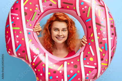 Shot of lovely cheerful red haired girl looks through swimring hole, has toothy smile, spends summer holidays on beach, likes swimming and tanning, enjoys awesome vacation. Seasonal rest concept photo