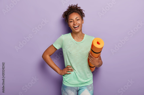 Fit positive Afro woman does fitness exercises and workout at home, holds orange karemat, keeps one hand on waist, wears casual t shirt and leggings, wants to loose weight, be healthy. Time to sport