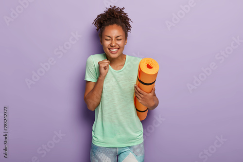 Bodycare and sport concept. Energized glad dark skinned woman clenches fist with joy, holds fitness mat, enjoys training session, works hard on getting healthier and fit, rejoices loosing weight