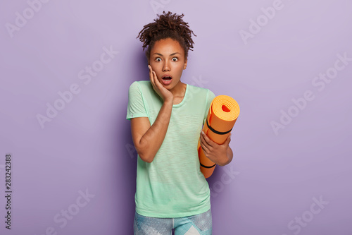 Keeping fit and healthy lifestyle concept. Shocked black woman with curly pony tail, holds orange fitness or yoga mat, surprised how much calories she needs to burn today, dressed in sportswear