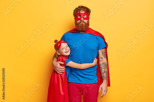 Happy childhood and fatherhood concept. Smiling small redhead female kid embraces with love father, wears bright superhero costume, come together on children parrty, pretend being supernatural.