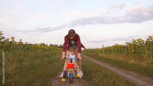 Mom teaches daughter to ride a bike on country road in a field of sunflowers. a small child learns to ride a bike. Mother plays with her little daughter. The concept of happy childhood.