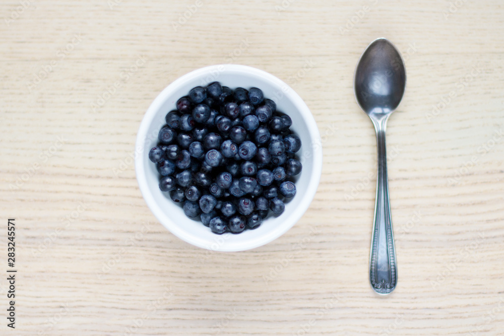 white ceramic bowl with blueberries, silver spoon on light wooden table white ceramic bowl with blueberries, silver spoon on light wooden table