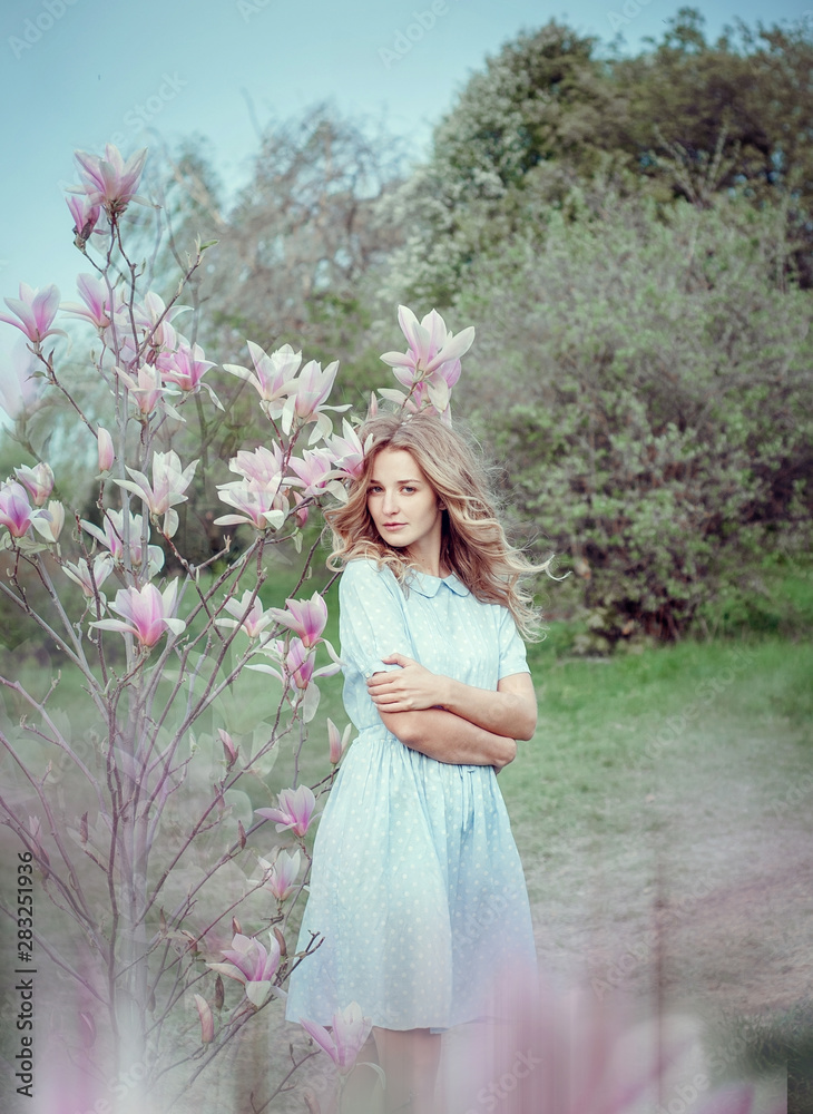 Beautiful happy young woman enjoying smell in a flowering spring garden.Beutiful tree magnolias,big flowers. Blonde with blue eyes and blue dress.