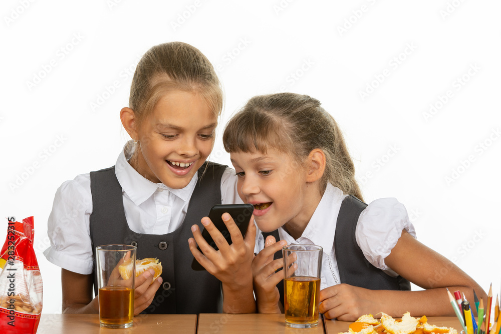 Two schoolgirls at a break watching a video on the phone, and eating liver and orange, drinking juice Two schoolgirls at a break watching a video on the phone, and eating liver and orange, drinking juice