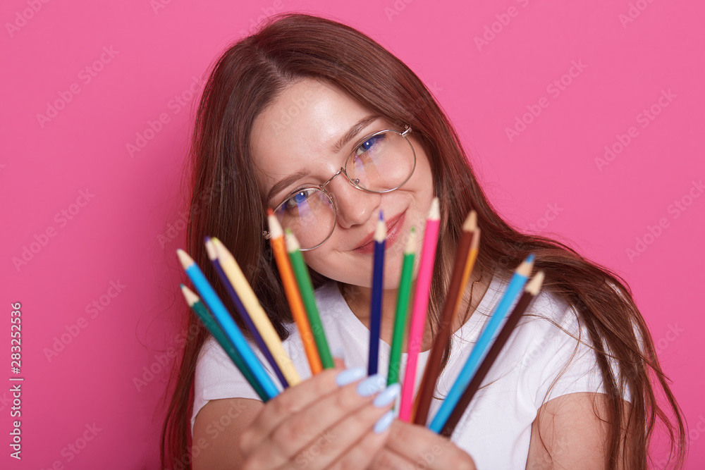 Indoor shot of young beautiful girl artist wearing white casual shirt and eyewear, model posing with colored pencils in her hands, charming female looks at camera, showig her pencils. Drowing concept. Indoor shot of young beautiful girl artist wearing white casual shirt and eyewear, model posing with colored pencils in her hands, charming female looks at camera, showig her pencils. Drowing concept.