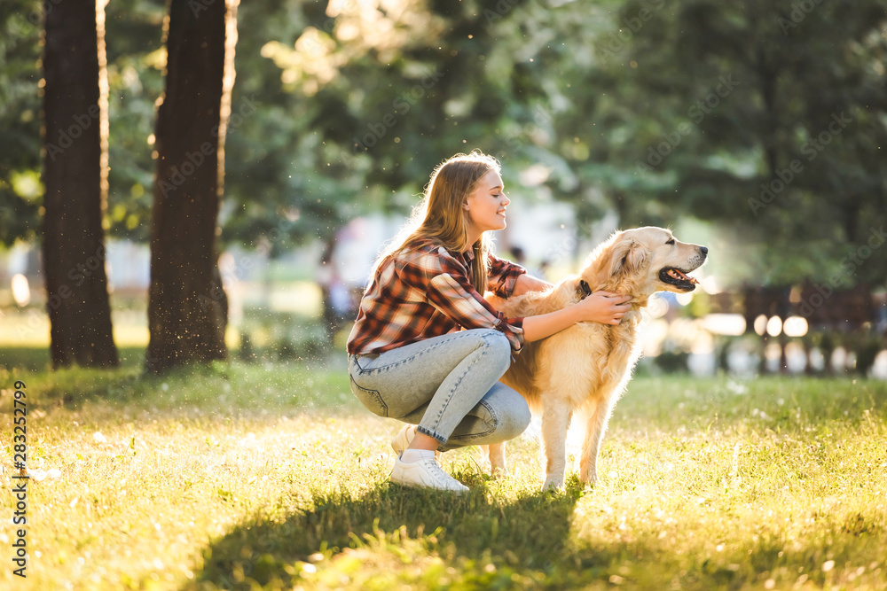 side view of beautiful girl in casual clothes petting golden retriever while sitting on meadow in sunlight side view of beautiful girl in casual clothes petting golden retriever while sitting on meadow in sunlight