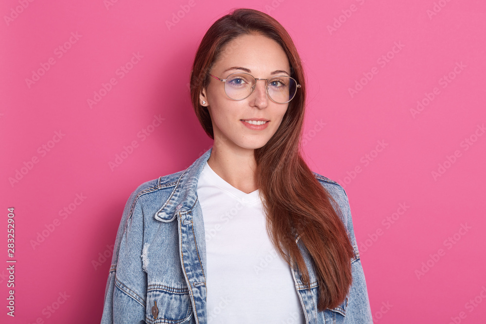 Indoor shot of pleasant looking woman, smiles positively, wearing optical glasses, white t shirt and denim jacket, being in hight spirt, model posing isolated over pink background. Copy space. Indoor shot of pleasant looking woman, smiles positively, wearing optical glasses, white t shirt and denim jacket, being in hight spirt, model posing isolated over pink background. Copy space.