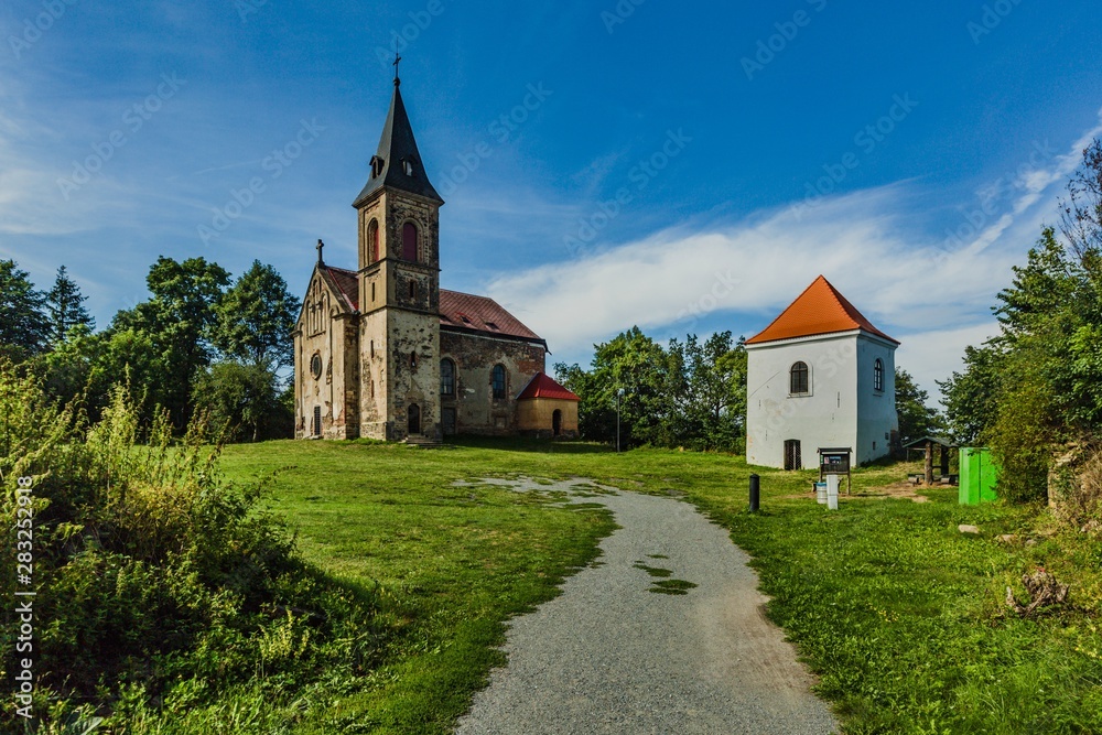 Krasikov, Kokasice / Czech Republic - August 9 2019: View of the church of Mary Magdalene and a bell tower. Bright sunny day with blue sky and white clouds. Gravel footh path leading to church.