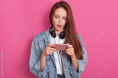 Image of stupefied woman staring at smart phone's screen with open mouth, watching horrible video in internet, wearing fashionable clothing, having beautiful long hair, isolated over rose background. photo