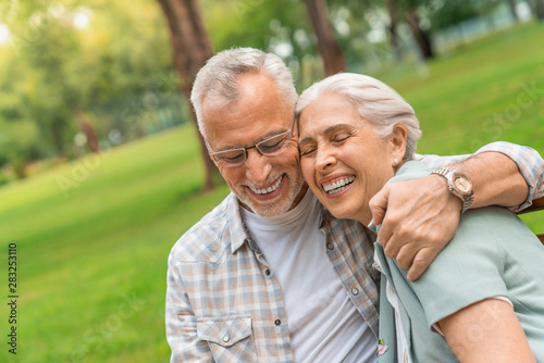 Smiling senior man and woman sitting on park bench hugging and laughing