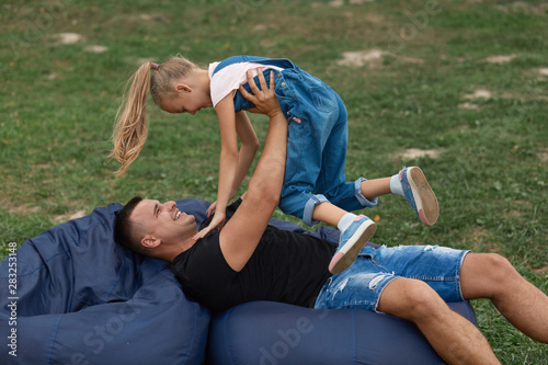 Happy father and doughter having fun and playing outdoors, sitting on frameless chair, spending time together in nature, spending holiday in park , dad and little girl wearing casual outfit.