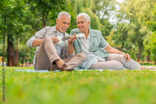 Senior man playing on ukulele to his wife on a picnic, seated on a blanket. The shot is in a city park during early autumn © InsideCreativeHouse