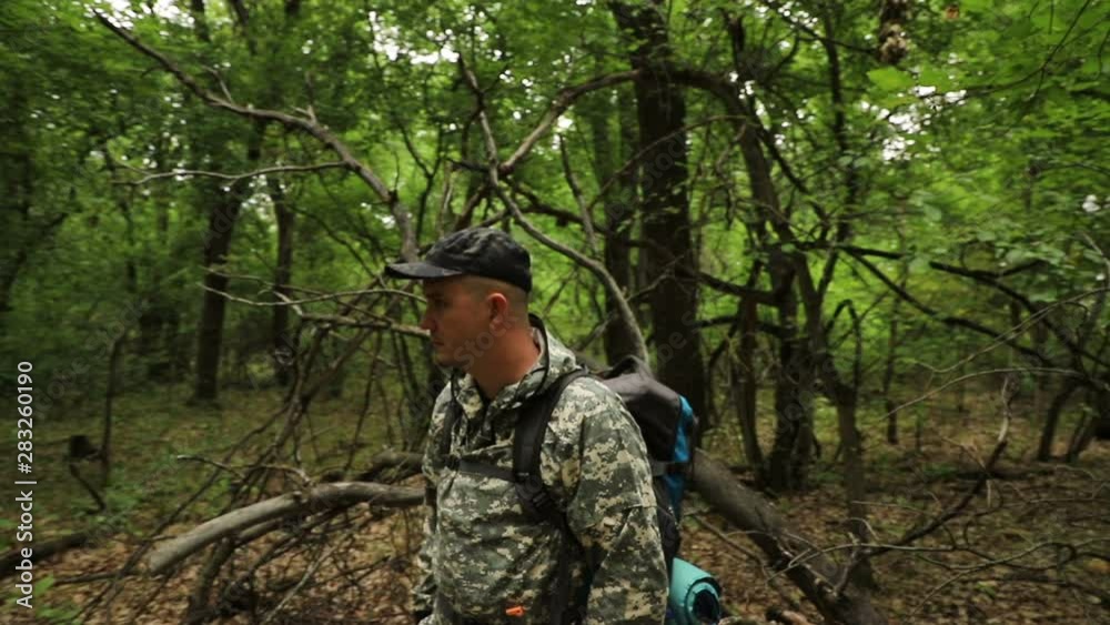 The camera moves around a caucasian handsome traveler in camouflage clothing, a cap and a backpack sitting on a log in a dense green forest, medium plan The camera moves around a caucasian handsome traveler in camouflage clothing, a cap and a backpack sitting on a log in a dense green forest, medium plan
