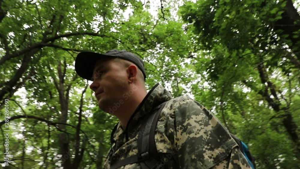 The camera moves around a caucasian handsome traveler in camouflage clothing, a cap and a backpack sitting on a log in a dense green forest, medium plan The camera moves around a caucasian handsome traveler in camouflage clothing, a cap and a backpack sitting on a log in a dense green forest, medium plan