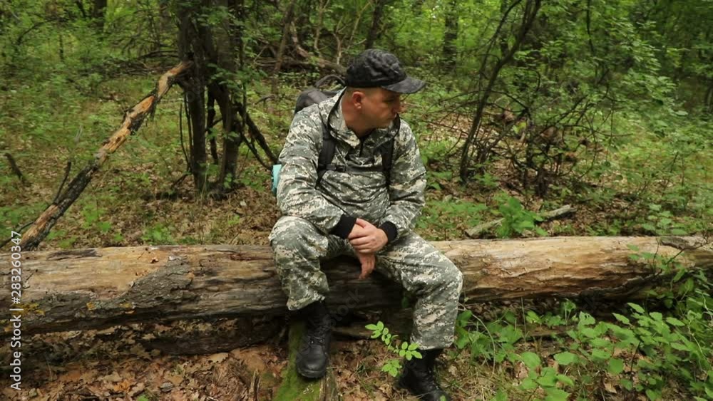 The camera moves around a caucasian handsome traveler in camouflage clothing, a cap and a backpack sitting on a log in a dense green forest, medium plan The camera moves around a caucasian handsome traveler in camouflage clothing, a cap and a backpack sitting on a log in a dense green forest, medium plan