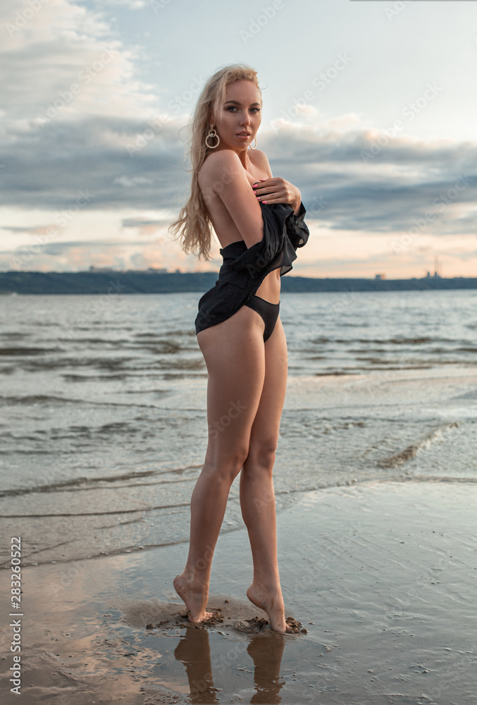 Blonde girl stands near the water at sunset. Hands clasped to chest