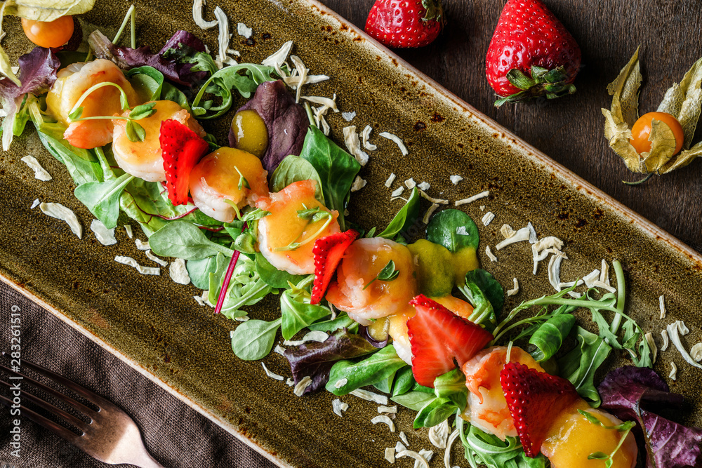 Healthy fresh salad with mix greens, strawberry, physalis and shrimp in plate over brown wooden background with linen napkin. Healthy food, clean eating, dieting, top view