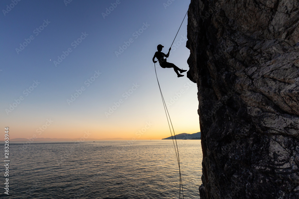 Silhouette of a Unrecognizable man rappelling down a steep cliff on the rocly ocean coast during a sunny summer sunset. Taken in Lighthouse Park, West Vancouver, British Columbia, Canada. - obrazy, fototapety, plakaty 