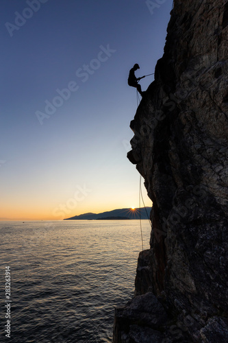 Silhouette of a Unrecognizable man rappelling down a steep cliff on the rocly ocean coast during a sunny summer sunset. Taken in Lighthouse Park, West Vancouver, British Columbia, Canada.