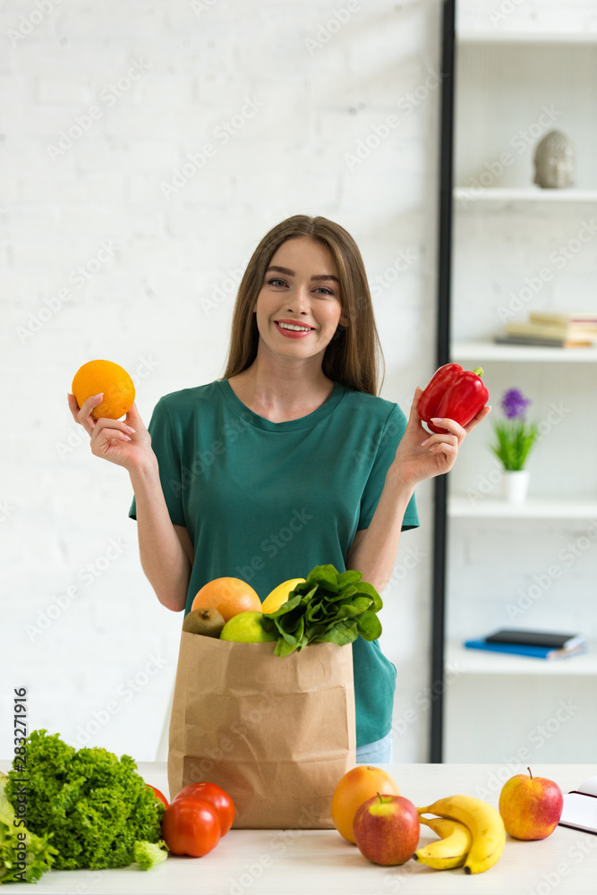 smiling young woman holding orange and bell pepper while standing near paper bag with fresh vegetables and fruits at home smiling young woman holding orange and bell pepper while standing near paper bag with fresh vegetables and fruits at home