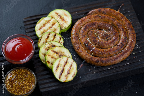 Grilled coiled sausage with zucchini slices and dipping sauces on a black wooden serving board, horizontal shot