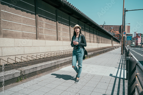 full length of girl traveler using smartphone on street in tokyo city. young woman walking along temple wall in urban and holding cellphone with online map app. lady backpacker travel japan in summer © PR Image Factory