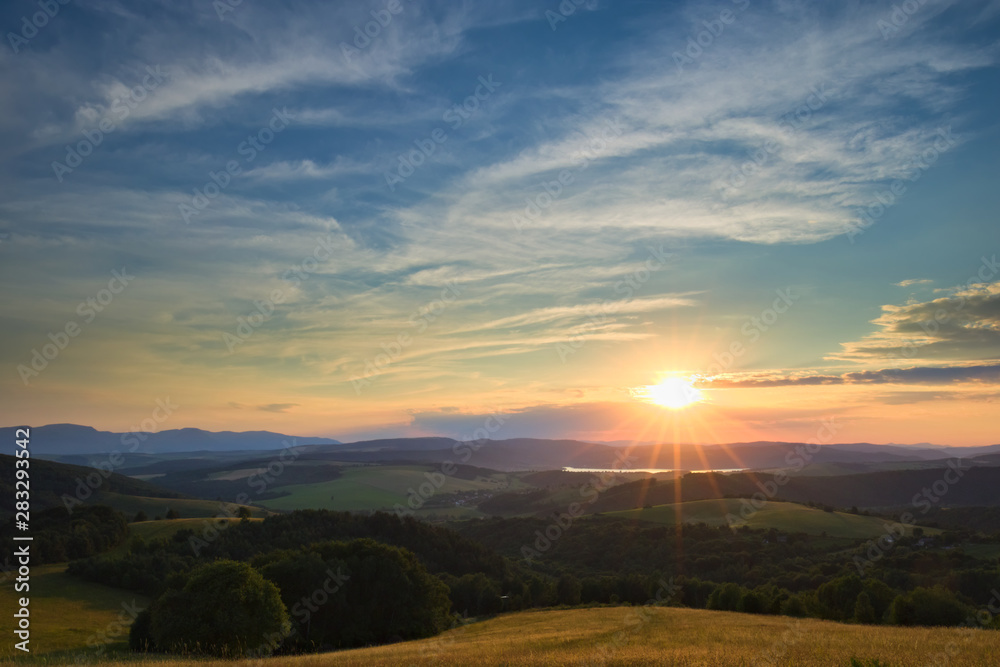 A calm sunset viewed from a hill with a meadow and a forest in the foreground and hills a mountain range and a lake in the background with nice clouds