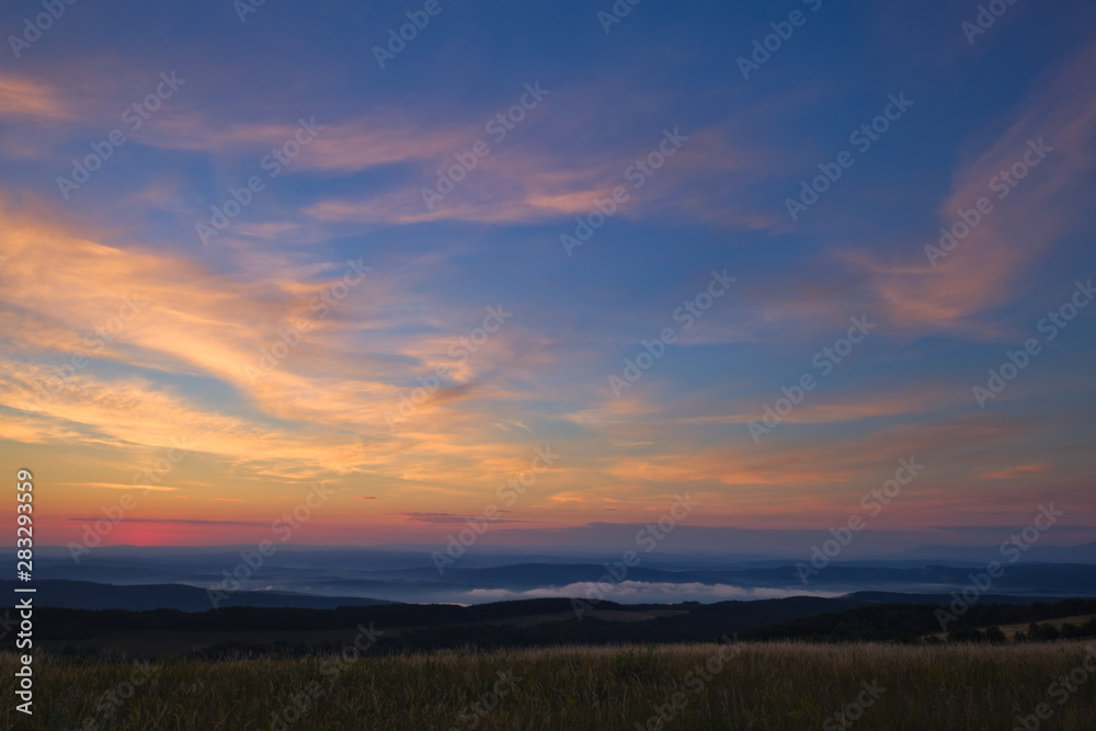 Slightly cloudy sky right before sunrise glowing with bright colors above hills, forests and meadows