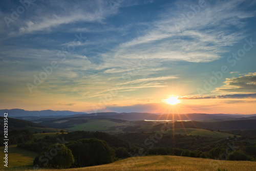 A calm sunset viewed from a hill with a meadow and a forest in the foreground and hills a mountain range and a lake in the background with nice clouds