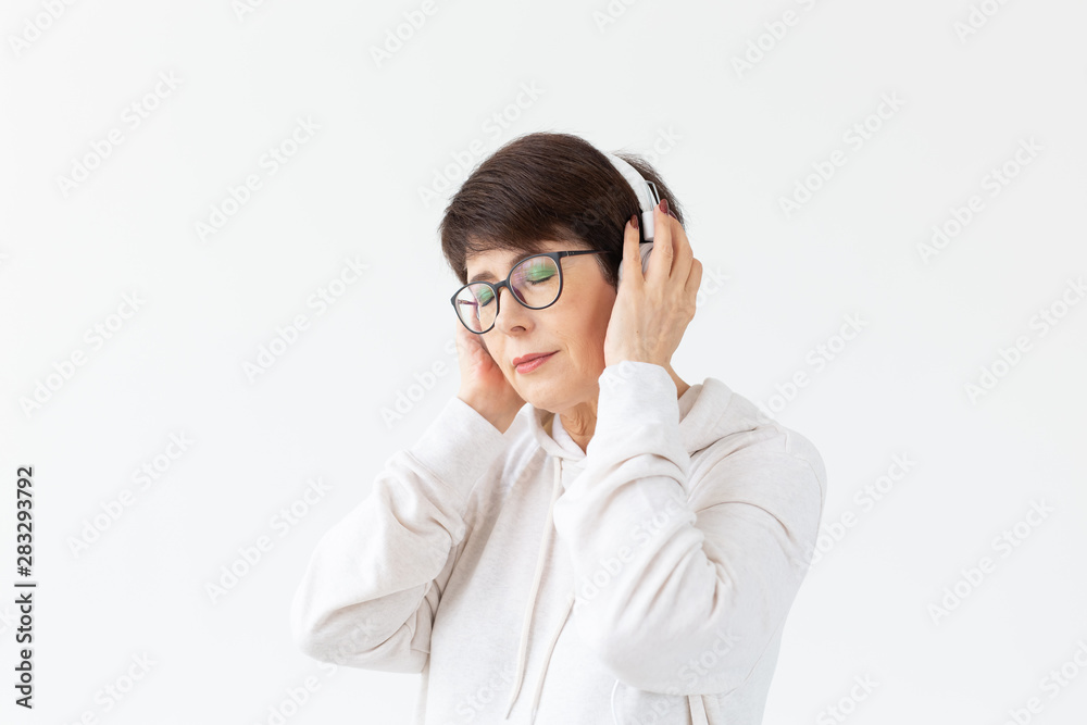 Pretty keen middle-aged woman in glasses and a white sweater listens to her favorite music with headphones on a white background with copy space. Online radio and music subscription concept.