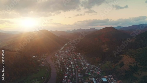 pictorial town at hills foot on winding river bank against sun rising from behind mountains in early autumn aerial view. Carpathian mountains, Ukraine beauty nature. Travel, summer holidays. 4K photo