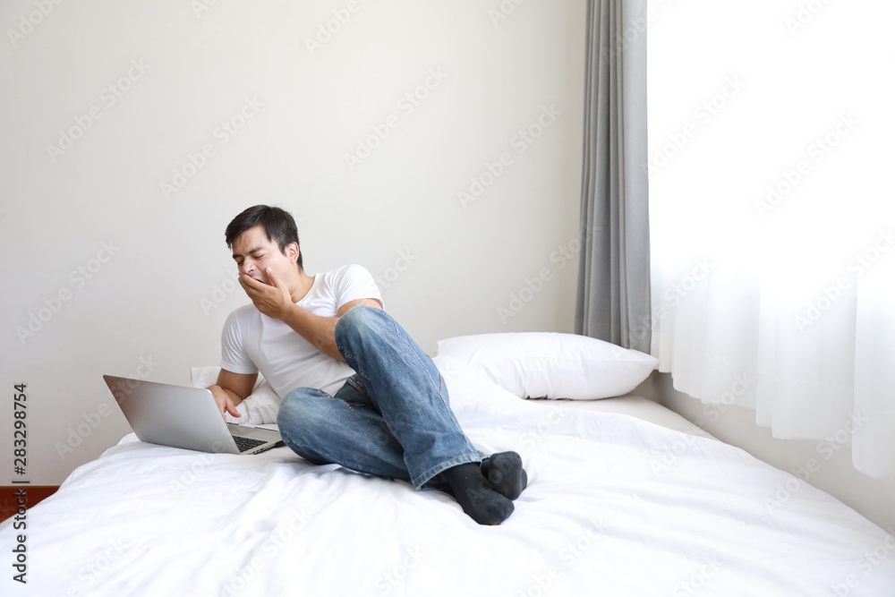 full shot of handsome caucasian in white shirt and blue jean using computer and yawning on bed in bedroom (work at home concept) full shot of handsome caucasian in white shirt and blue jean using computer and yawning on bed in bedroom (work at home concept)