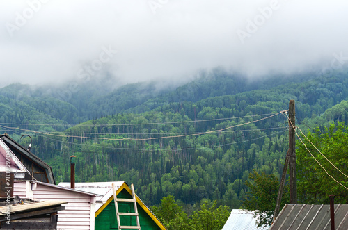 Rooftops of Traditional Houses, Metal Pipe Chimneys, A Ladder, Old Power Pole, Wires, Garden Trees On A Misty Summer Morning With Mountain Slopes Of Mixed Forest And Low Flowing Clouds In Background.