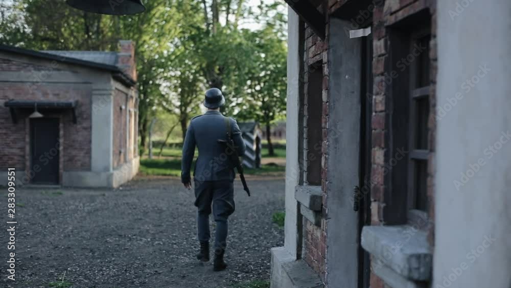 Back view of a young man in a German WW2 uniform walking outside. Red brick historical buildings on the background Back view of a young man in a German WW2 uniform walking outside. Red brick historical buildings on the background