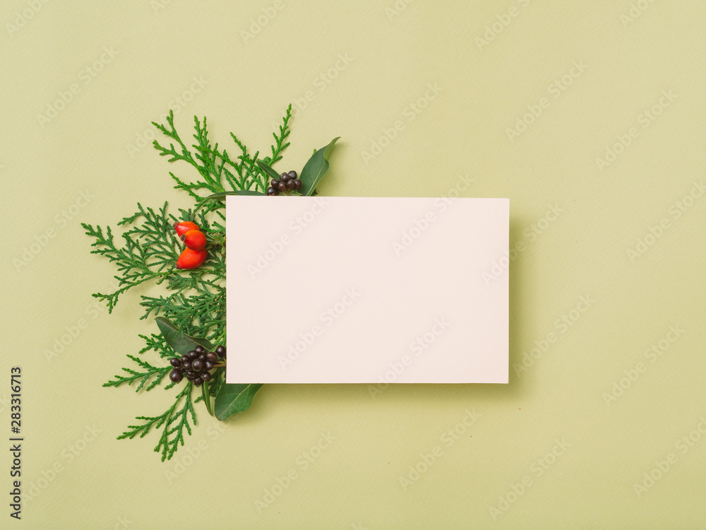 Festive greeting card. White mockup paper sheet with green juniper sprig and fall berries decor on olive background. Copy space.