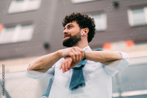 Successful man with talking via ear beands leaning on the railing next to modern glass buildings resting outdoors.