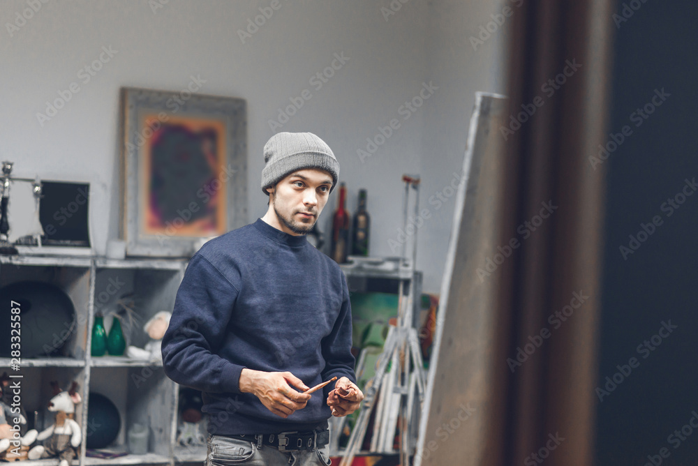 Portrait of an artist student with a brush in his hand in a cozy art studio near an easel with a canvas. Man studies painting in an art studio. Learning painting. Portrait of an artist student with a brush in his hand in a cozy art studio near an easel with a canvas. Man studies painting in an art studio. Learning painting.