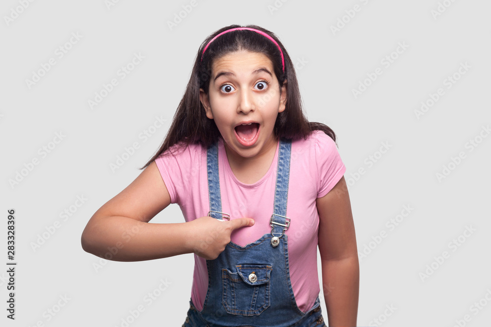 Φωτογραφία who? me? Portrait of shocked brunette girl in casual pink t-shirt and blue overalls standing, pointing herself, looking at camera with surprised face