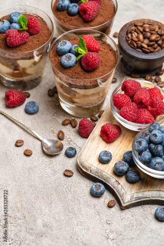 Classic tiramisu dessert with blueberries and raspberries in a glass and bowls with berries on concrete background