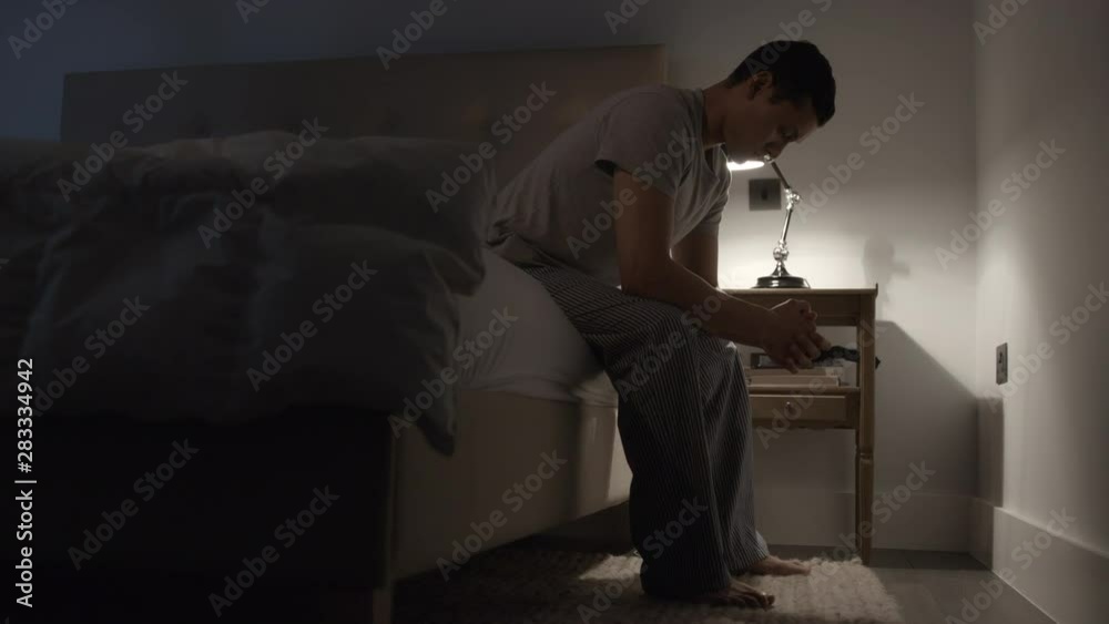 Stressed and unhappy man sitting on the edge of a bed at night. His thoughts troubling his mind Stressed and unhappy man sitting on the edge of a bed at night. His thoughts troubling his mind