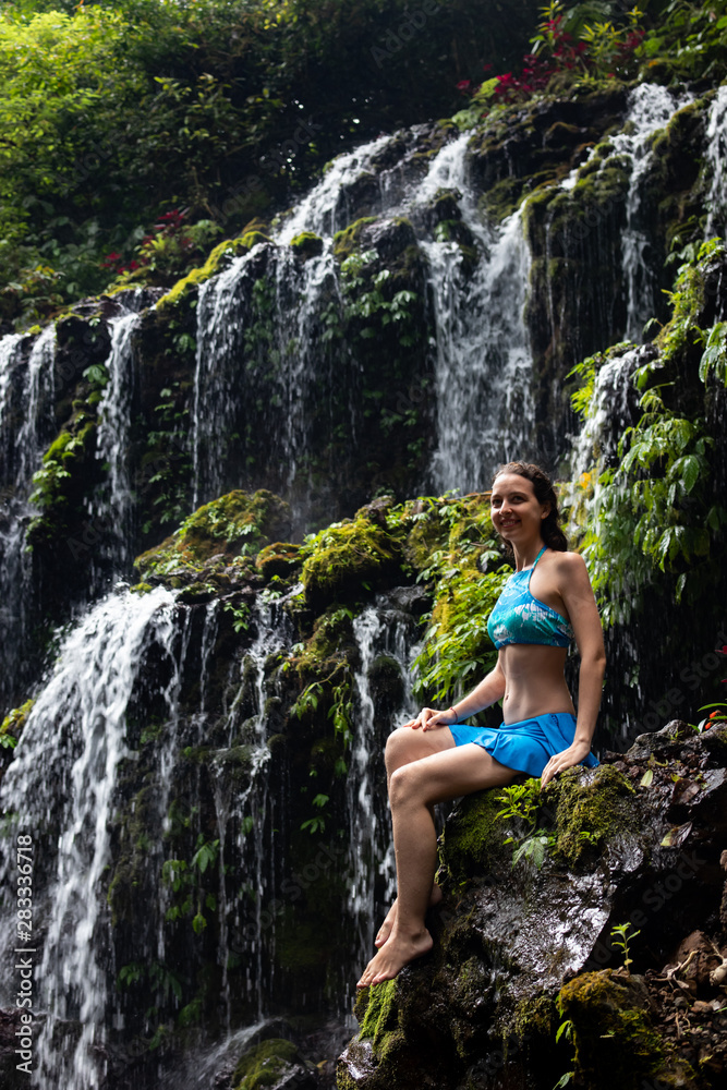 Travel lifestyle. Young traveler woman sitting at waterfall in tropical forest. Banyu Wana Amertha waterfall Wanagiri, Bali, Indonesia. Travel lifestyle. Young traveler woman sitting at waterfall in tropical forest. Banyu Wana Amertha waterfall Wanagiri, Bali, Indonesia.
