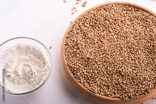 Wheat seeds grains in wooden bowl near with wheat flour in glass bowl, heap of grains, angle view, isolated on white background, copy space