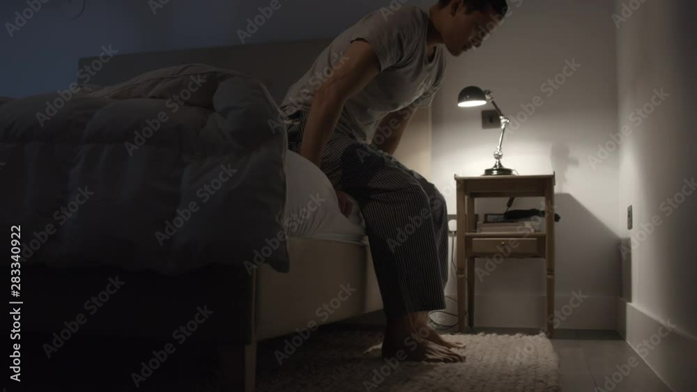 Young man getting out and sitting on the edge of the bed at night suffering with anxiety, stress and depression Young man getting out and sitting on the edge of the bed at night suffering with anxiety, stress and depression