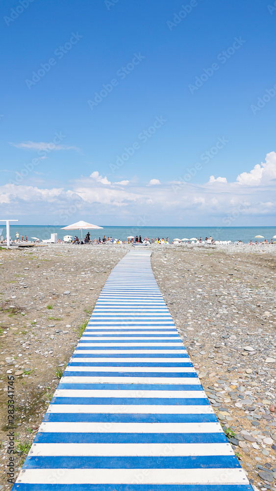 Wooden blue and white striped Board Path Leading through pebbles to the Beach. Blue sky, sea, vacation, travel.