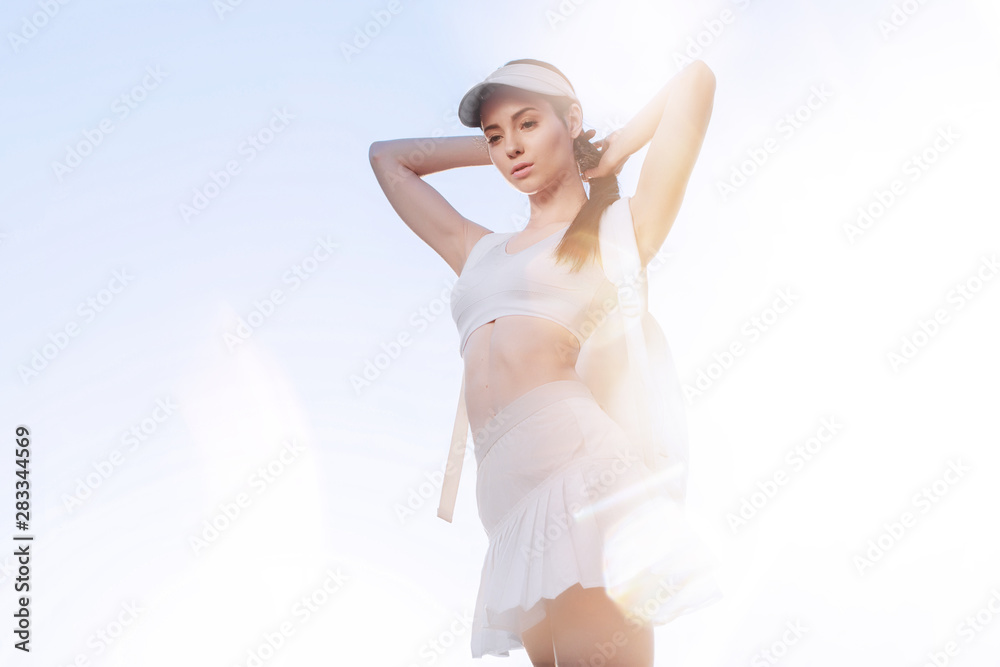 Naklejka premium Sports woman posing in fashion sportswear on sunny sky background. Beautiful slim girl in trendy white skirt, top, cap and backpack. 