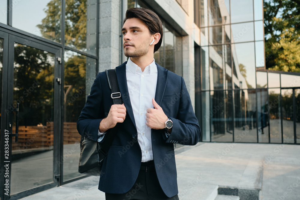 Young handsome bearded brunette man in white shirt and classic suit with wireless earphones and backpack on shoulder dreamily looking aside with building on background Young handsome bearded brunette man in white shirt and classic suit with wireless earphones and backpack on shoulder dreamily looking aside with building on background
